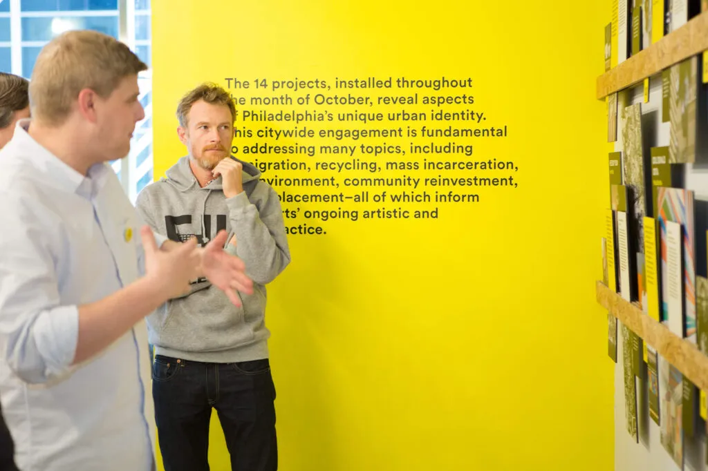 Artists discussing exhibition in front of a yellow wall for city-wide art exhibition for Mural Arts by J2