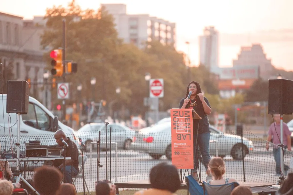 African American woman speaking at a podium with orange and black typographic logo with city in the background