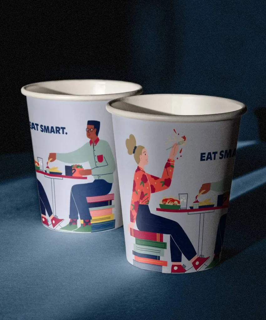 Two paper coffee cups wrapped in vibrant and quirky brand illustrations of students in dining hall using books as seats