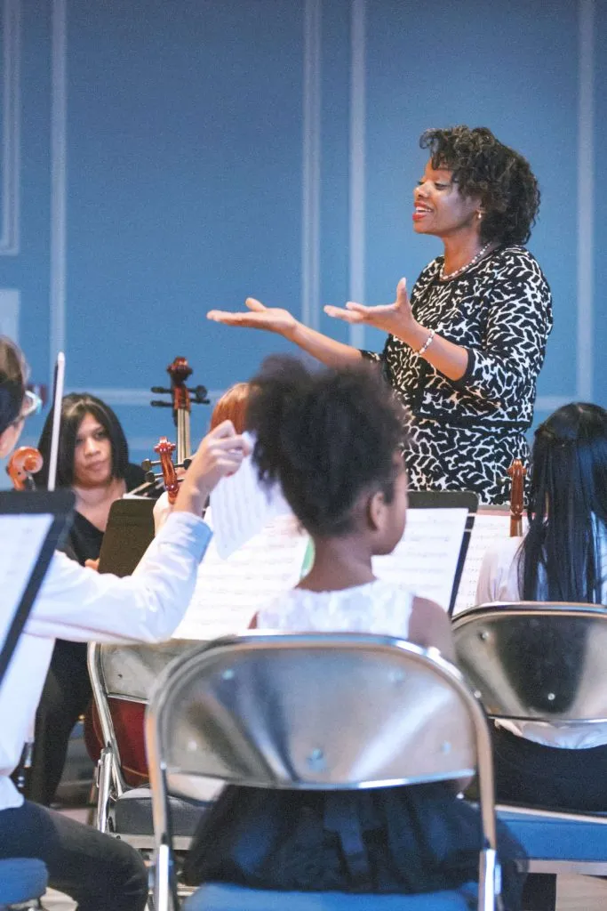 African American woman music teacher addressing class of students holding string instruments