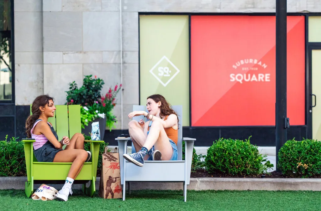 two teenage girls sitting in courtyard on colorful chairs with bold logo graphics on windows in background