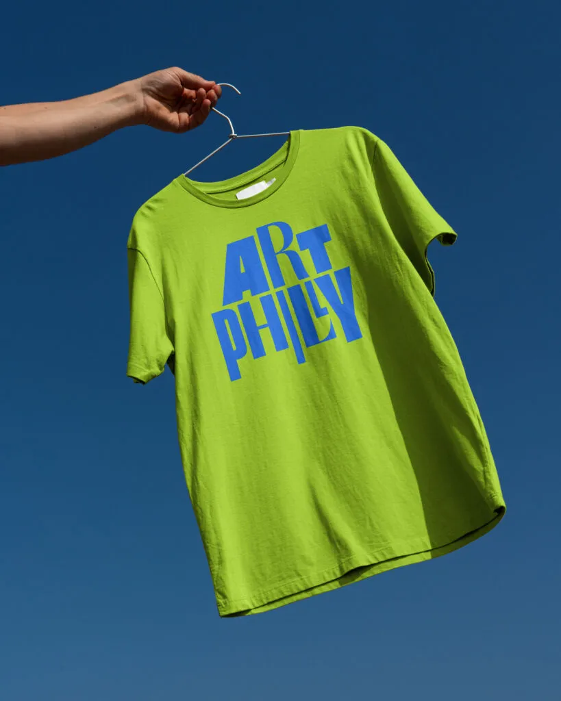 Bright green tshirt on a hanger featuring ArtPhilly arts festival logo by J2.