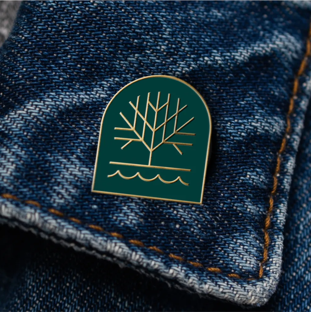 Enamel pin with tree and water logo attached to a jean jacket.