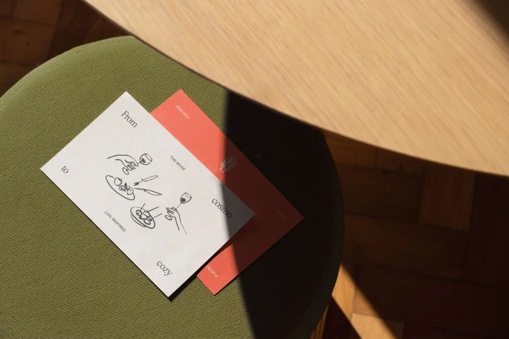 business cards featuring custom illustrations of two residents enjoying a meal. The cards are placed on a green fabric chair in front of a light wood dining table. The Muse branding by J2